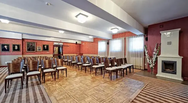 Elegancka sala konferencyjna w Lanckorona Hotel Modrzewiówka, z rzędami krzeseł, portretami na ścianach i ozdobnym piecem.