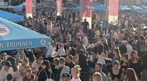 Menschenmenge feiert beim Bamberger Weinfest. Eine Bühne mit Musikern, Fahnen und Zelten. Viele Besucher sitzen an Tischen, trinken und genießen die Atmosphäre.