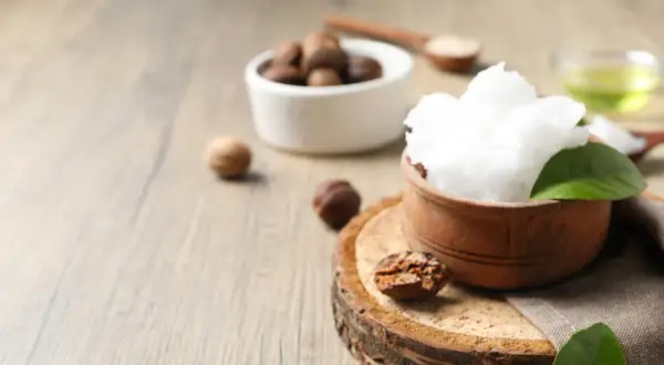 Un tazón de manteca de karité pura y cremosa, rodeado de nueces de karité y hojas verdes, sobre una superficie de madera rústica.