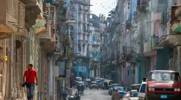 Photographie de rue d'une rue cubaine animée, avec des bâtiments colorés et délabrés, des voitures anciennes et un homme marchant seul.