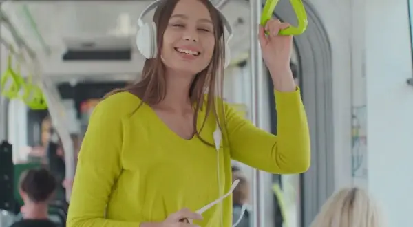 Uśmiechnięta dziewczyna w słuchawkach słucha podcastów po angielsku w tramwaju, trzymając się poręczy.