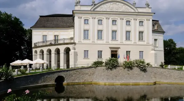 Pałac w Łącku, biała, okazała budowla z arkadowym tarasem, otoczona fosą i zielenią.
