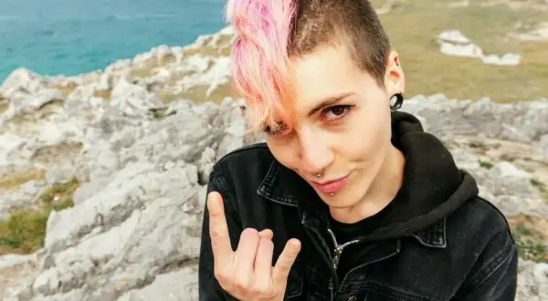 Dziewczyna z różowymi włosami i kolczykami, w kurtce dżinsowej, pozuje na tle skał i morza, pokazując znak rocka. Jej styl to czysty **punk**.