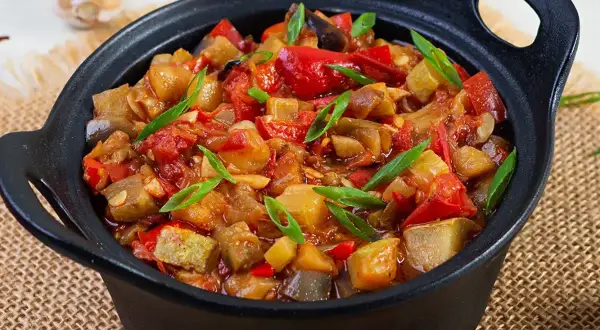 Un plat de ratatouille colorée, avec des morceaux d'aubergine, de courgette, de poivron rouge et de tomate, garni de ciboule fraîche. Une recette savoureuse.