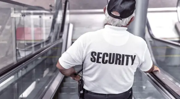 Pracownik ochrony w białej koszulce z napisem "SECURITY" stoi na ruchomych schodach, czuwając nad bezpieczeństwem.