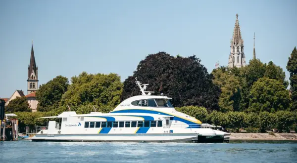 Schnelles Katamaran-Schiff auf dem Bodensee, mit Kirchtürmen von Friedrichshafen und Konstanz im Hintergrund.