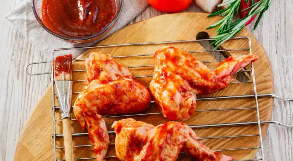 Skrzydełka kurczaka w aromatycznej marynacie na grilla, gotowe do pieczenia. Obok miseczka sosu i pędzelek.