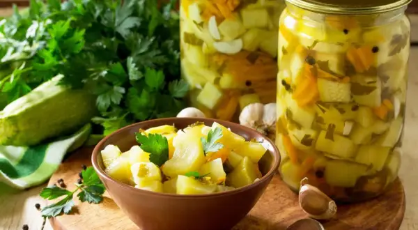 Słoiki z cukinią w zalewie słodko-kwaśnej z curry, miseczka z marynowaną cukinią, świeża cukinia, czosnek i natka pietruszki.