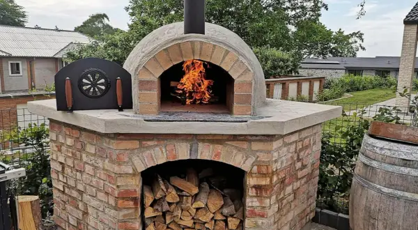 En vacker, murad pizzaugn med eld som sprakar i öppningen. Ved ligger staplad under ugnen, redo för nästa pizzakväll.