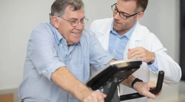 Un homme âgé sourit en effectuant un **test d'effort vélo**, supervisé par un médecin en blouse blanche.