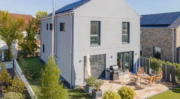 Ein modernes, schmales Haus mit Solaranlage auf dem Dach und einer Terrasse mit Grill und Sitzmöbeln.