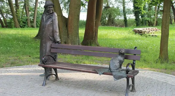 Pomnik mężczyzny w kapeluszu i kota na ławce w parku.