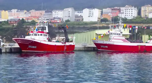 Dos barcos pesqueros rojos y blancos, listos para zarpar, adornados con banderas de gala. En el fondo, un pueblo costero con edificios de colores, evocando la tradición de la virxe da mariña.