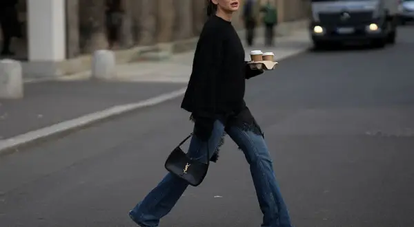 Kobieta w jeansach bootcut i czarnym swetrze z koronkowymi wstawkami niesie kawę. Stylowe jeansy bootcut to podstawa wielu casualowych stylizacji.
