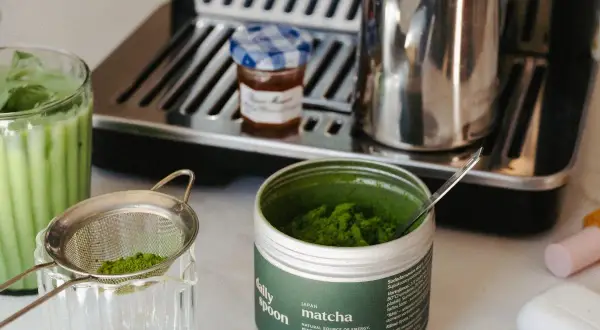 Puszka z zielonym proszkiem matcha, sitko z proszkiem, szklanka z napojem i ekspres do kawy.