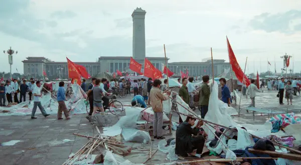 Ludzie na placu Tiananmen, czerwone flagi powiewają, budynki w tle, chaos i prowizoryczne namioty.