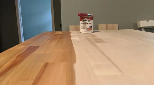 A can of Watco Butcher Block Oil & Finish sits on a wooden surface, with one half of the butcher block already stained and the other half awaiting treatment.