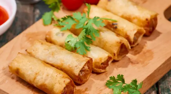 Chrupiące spring rolls na desce, ozdobione kolendrą i pomidorkami. Idealne na przekąskę.