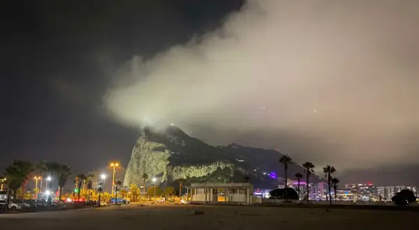 Nocny widok na Gibraltar, z chmurami otulającymi skałę. W oddali widać oświetlone miasto i palmy.