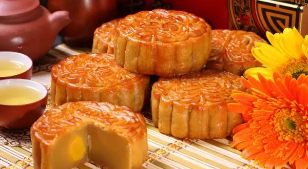 Ciastka księżycowe i herbata na święto środka jesieni. Złote ciastka z nadzieniem, ozdobione wzorami, obok filiżanek z herbatą i kwiatów.
