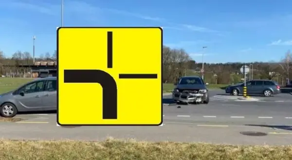 Znak drogowy informuje, kto ma pierwszeństwo na skrzyżowaniu równorzędnym. Widać uszkodzone auto po wypadku.