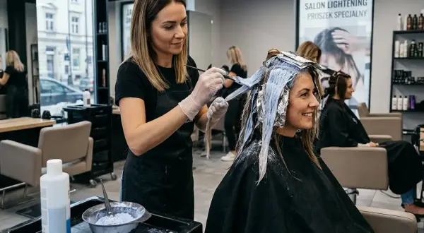 Fryzjerka wykonuje rozjaśnianie włosów klientce, nakładając farbę za pomocą pędzla. W tle widać inne klientki i półki z kosmetykami.