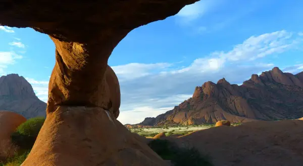 Felsformationen der Spitzkoppe in Namibia, mit einem Felsentor im Vordergrund und zerklüfteten Bergen am Horizont unter blauem Himmel.