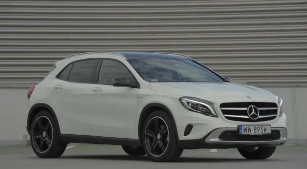 Biały Mercedes GLA na tle szarej, falistej ściany. Zbliżenie na przód auta, widoczne logo i tablica rejestracyjna. Idealny do miasta, poznaj jego mercedes gla wymiary.