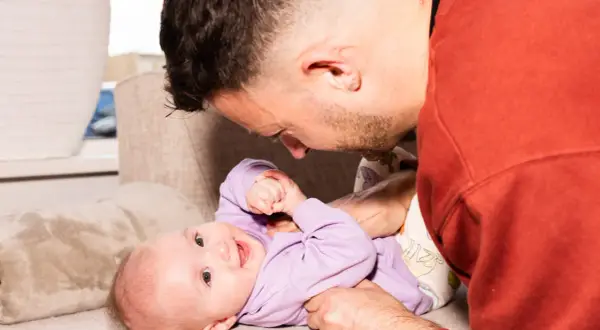 Ein Vater spielt liebevoll mit seinem 4. sprung baby auf einer Couch. Das Baby trägt einen lila Pullover und lächelt.