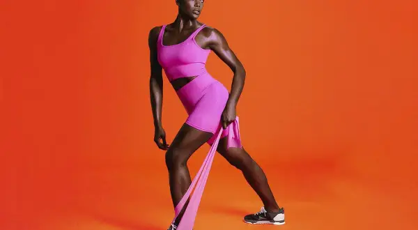Femme athlétique en tenue rose vif, effectuant un exercice avec élastique sur fond orange vif.