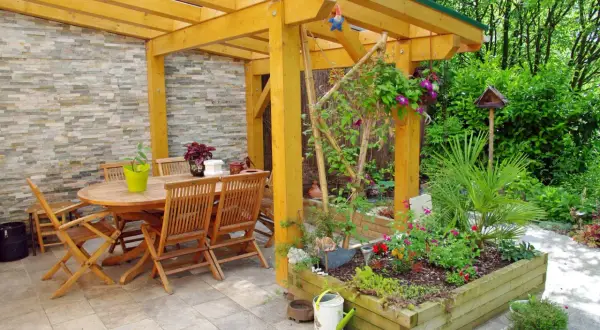 Un pergolato terrazzo fai da te accoglie un tavolo in legno e sedie, circondato da aiuole fiorite e vegetazione lussureggiante.