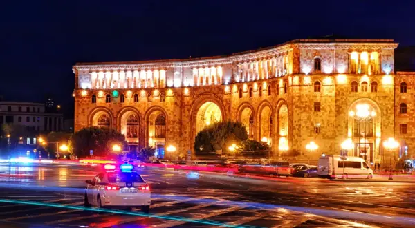 Nocna panorama placu w Erywaniu, największym mieście Armenii. Oświetlony budynek z arkadami, na ulicy widać ślady świateł samochodów i radiowóz.