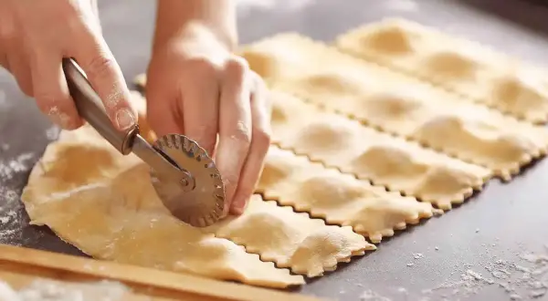 Préparation d'une recette de ravioli maison. Des mains découpent la pâte avec une roulette dentelée, formant des carrés prêts à être farcis.