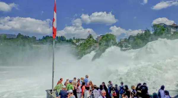 Menschen bestaunen den tosenden Rheinfall. Eine Schweizer Fahne weht im Wind. Der **Rheinfall Eintritt** ist ein beeindruckendes Naturschauspiel.