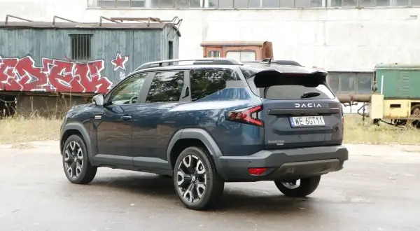 Ciemnoniebieska Dacia Bigster, model hybrydowy, zaparkowana na tle opuszczonej fabryki z wagonami kolejowymi pokrytymi graffiti.