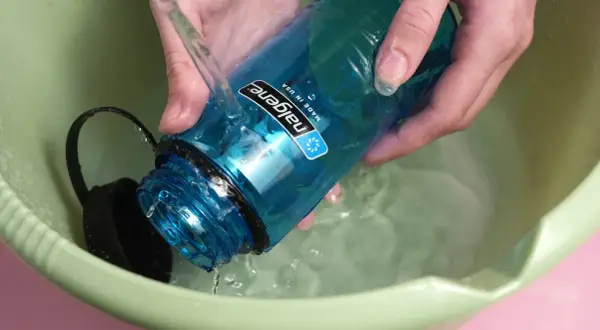 Hände spülen eine blaue Nalgene-Flasche mit kochendem Wasser in einer grünen Schüssel.