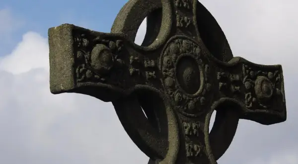 Kamienny krzyż celtycki z misternymi zdobieniami na tle błękitnego nieba z chmurami.
