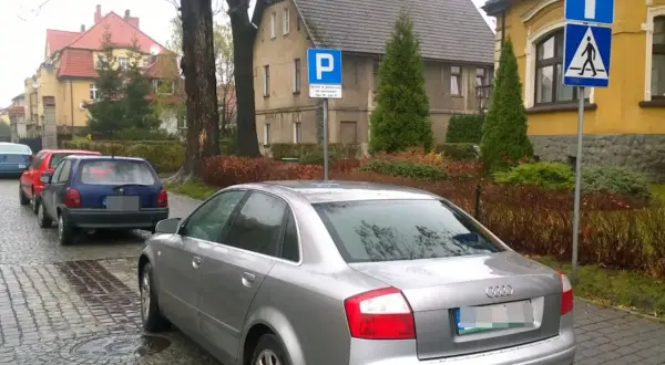 Samochody stoją na ulicy, a obok znak informuje o parkowaniu za przejściem dla pieszych.