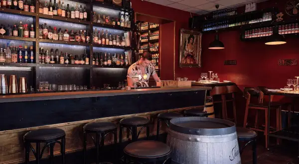 En bar med hyllor fyllda med flaskor, en bardisk med pallar och en bartender. En perfekt plats för vinbarer Stockholm.
