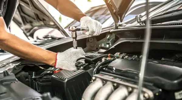 Mechanik w białych rękawiczkach dokręca śrubę kluczem nasadowym. Silnik samochodu jest otwarty, co sugeruje przegląd samochodu po terminie.