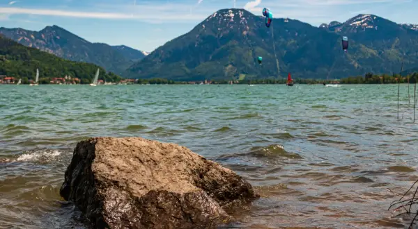 Kitesurfer tanzen über das Wasser am Bad Wiessee, während Segelboote und Berge im Hintergrund ruhen.