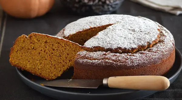Un delicioso bizcocho de calabaza espolvoreado con azúcar glas, con una rebanada cortada y un cuchillo listo para servir.