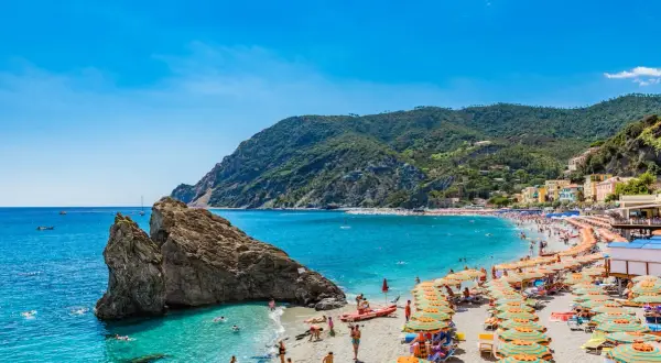 Spiaggia affollata sulla Riviera di Levante, con ombrelloni colorati, gente che nuota e montagne verdi sullo sfondo.