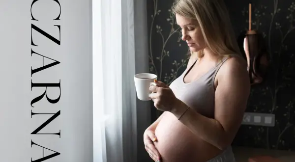 Ciężarna kobieta z kubkiem czarnej herbaty w dłoni, spogląda przez okno. Delikatne światło podkreśla jej brzuszek.