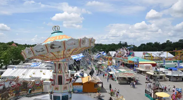 Buntes Treiben auf dem Volksfest Ingolstadt mit Kettenkarussell, Fahrgeschäften und Buden unter blauem Himmel mit Wolken.