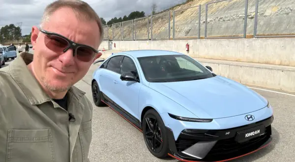 Mężczyzna w okularach przeciwsłonecznych pozuje do selfie z błękitnym Hyundaiem IONIQ 6 N na torze wyścigowym.