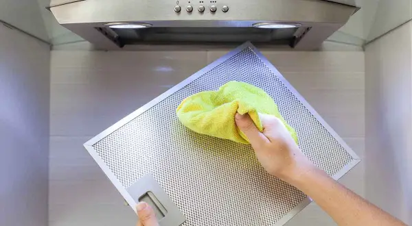 Mano pulisce il filtro metallico della cappa con un panno giallo. Impara come pulire i filtri della cappa per una cucina più sana.