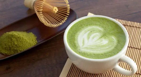 Tazza di matcha latte con schiuma decorata a forma di cuore, accanto a polvere di tè verde e frusta tradizionale.
