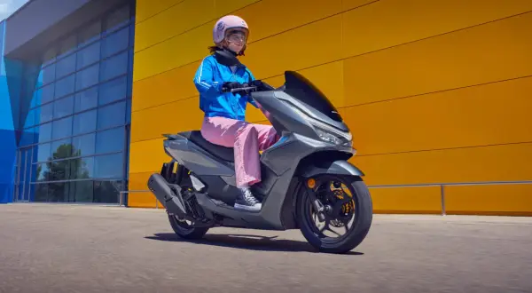Kobieta w różowych spodniach i niebieskiej kurtce jedzie na szarym skuterze Honda PCX 125. Wady tego modelu mogą dotyczyć np. zawieszenia.