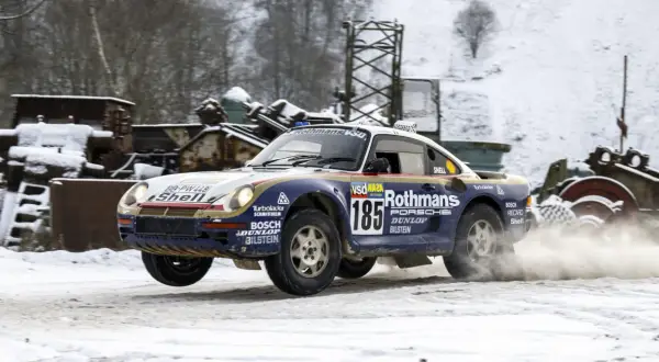 Porsche 959 w barwach Rothmans, z numerem 185, pokonuje śnieżny odcinek rajdu, wzbijając tumany śniegu.